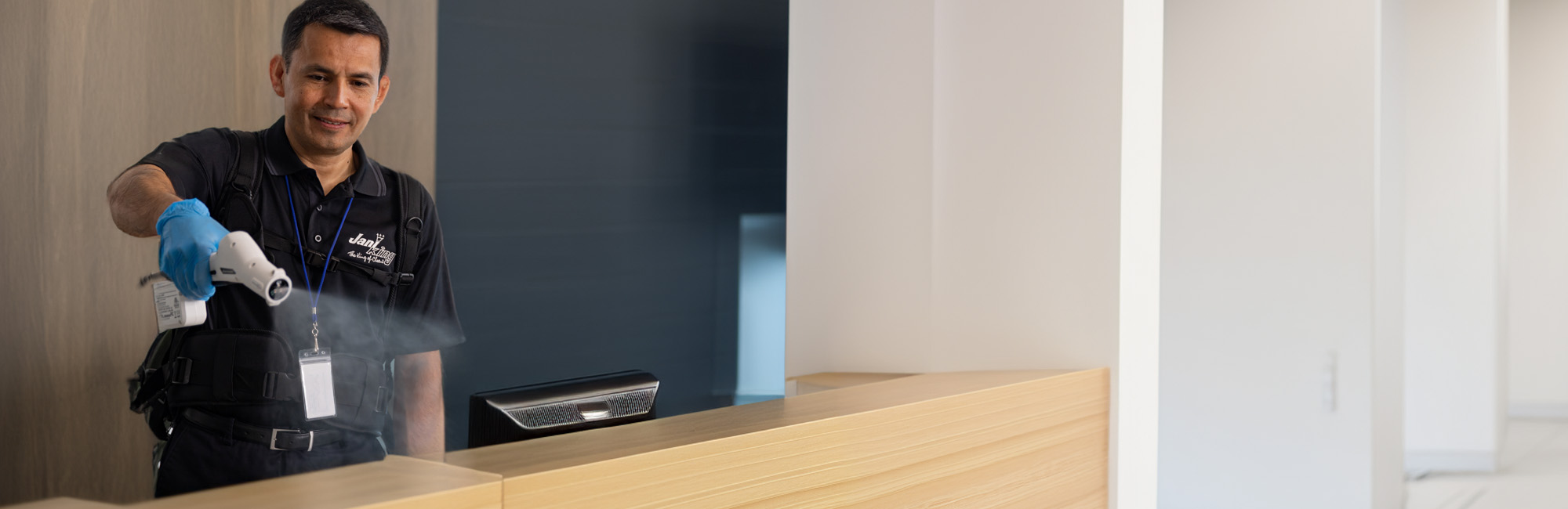 A person wearing a uniform and blue gloves is meticulously disinfecting a wooden counter with a spray device, embodying the essence of summer cleaning services in a clean, modern interior space. The background features light-colored walls and a minimalistic design.