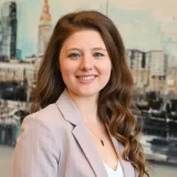 A woman with long, wavy brown hair is smiling. She is wearing a light-colored blazer over a white top. The background features a blurred cityscape with buildings. A woman with long, wavy brown hair is smiling. She is wearing a light-colored blazer over a white top.
